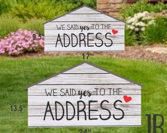 May include: Two wooden signs with a house shape and the text "We said yes to the address" with a red heart. The larger sign is 24 inches wide and 13.5 inches tall. The smaller sign is 17 inches wide and 9.5 inches tall. The signs are on a green lawn.