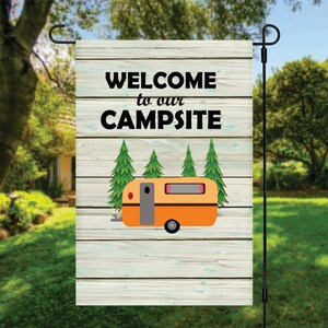 May include: Welcome to our campsite garden flag with a woodgrain background, featuring a yellow camper with a gray door and three green pine trees.