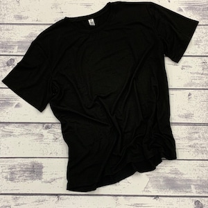 May include: A solid black crew neck t-shirt. The short-sleeved shirt is made of a soft material and is laying on a light-colored wooden surface. The tag on the shirt reads "JB".