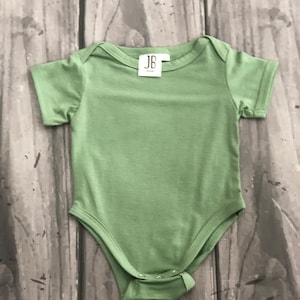 May include: A light green baby onesie with short sleeves and a round neckline. The onesie has a snap closure at the bottom and a small tag near the neckline with the letters "JB". The garment is made of a soft material.