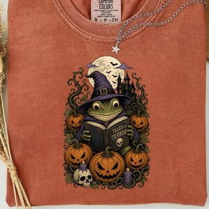 Puede incluir: Una camiseta color óxido con un estampado gráfico de una rana verde con un sombrero de bruja morado leyendo un libro titulado Tales of Terror. El diseño incluye calabazas naranjas, una calavera, velas moradas y una casa encantada.
