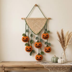 May include: A wall hanging featuring a triangular beige crochet piece suspended from a wooden dowel by a green cord. Six small orange crocheted pumpkins with black stitched jack-o'-lantern faces and green leaves hang from the base on green cords.