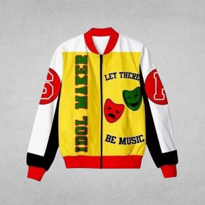 May include: A zip-up bomber jacket featuring a yellow front panel with the text IDOL MAKER and LET THERE BE MUSIC, accompanied by red and green theatrical masks. The sleeves are white with black accents and red cuffs, featuring red circular patches.