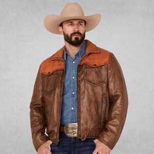 May include: A beige cowboy hat, a blue button-down shirt, and a brown leather jacket with a contrasting tan leather yoke across the shoulders. The jacket features a front zipper closure, two chest pockets, and a silver belt buckle at the waist.