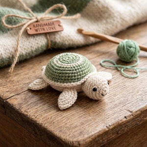 Puede incluir: Un pequeño juguete de tortuga tejido a ganchillo con cuerpo color crema y un caparazón con patrón en verde salvia y crema. La tortuga tiene ojos de cuentas negras y una sonrisa bordada. Una etiqueta de madera con HANDMADE WITH LOVE está atada.