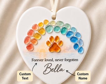 May include: A white heart-shaped ceramic ornament hanging from a cream ribbon. It features a rainbow of colorful glass pebbles, a central orange paw print, a sunset illustration, and the text Forever loved, never forgotten and Bella.