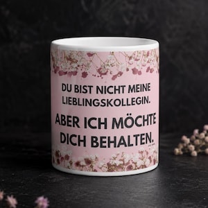 Könnte beinhalten: Ein weißer Keramikbecher mit rosa Hintergrund und einer floralen Bordüre aus kleinen weißen und rosa Blüten. Der Becher zeigt den schwarzen deutschen Text: DU BIST NICHT MEINE LIEBLINGSKOLLEGIN. ABER ICH MÖCHTE DICH BEHALTEN.