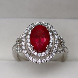 Puede incluir: Un anillo de tono plateado con una gran piedra preciosa de color rojo intenso y corte ovalado en el centro. La piedra central está rodeada por un doble halo de pequeñas piedras claras de corte redondo. El aro también tiene piedras claras.