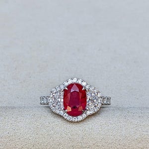 Könnte beinhalten: Ein silberfarbener Ring mit einem großen, oval geschliffenen, tiefroten Rubin in der Mitte. Der Rubin ist von einem Kranz aus kleinen, klaren, rund geschliffenen weißen Steinen umgeben, mit weiteren kleinen weißen Steinen entlang der Ringschiene.