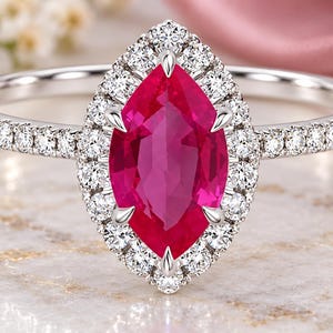 Op de afbeelding: Een zilverkleurige ring met een centrale marquise-geslepen felrode edelsteen, omringd door een halo van kleine ronde witte steentjes. De slanke ringband is aan beide zijden bezet met een rij kleine ronde witte steentjes bij de zetting.