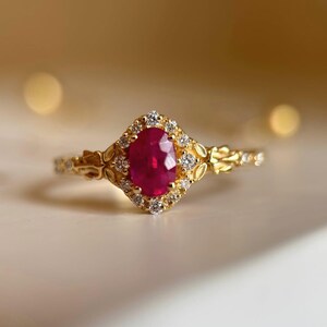 Puede incluir: Un anillo de oro con un rubí rojo central de corte ovalado rodeado por un halo de pequeños diamantes blancos redondos. La banda presenta intrincados detalles metálicos en forma de hoja que se extienden desde el engaste a lo largo del aro.
