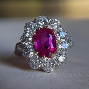 Puede incluir: Un anillo con una piedra preciosa central de corte ovalado en color rojo rosado vibrante, rodeada por un halo de diez diamantes blancos redondos. Las piedras están montadas en una banda metálica, con la piedra central sujeta por pequeñas garras doradas.