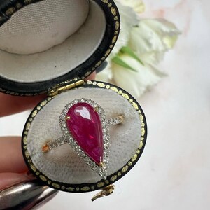 May include: A pear-shaped ruby ring set in a gold band with a halo of small white diamonds. The ring is displayed inside an oval-shaped black velvet jewellery box with a cream-coloured interior lining, showcasing the deep red gemstone clearly.