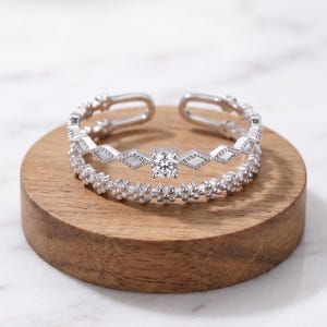 May include: A silver-toned open ring featuring two distinct bands. The top band displays a series of geometric diamond-shaped links with a single clear round stone at the centre. The bottom band consists of a continuous row of small floral shapes.