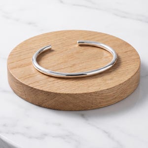 Op de afbeelding: Een gepolijste zilveren armband met een glad, afgerond oppervlak en een eenvoudig open ontwerp. De armband heeft een continue, uniforme dikte door de hele cirkelvormige structuur en toont een reflecterende metalen afwerking met strakke lijnen.