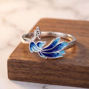 May include: A silver-toned metal ring featuring a decorative element shaped like a bird with a long, flowing tail. The tail is filled with enamel in shades of light blue, medium blue, and deep navy blue, creating a smooth, glossy finish.