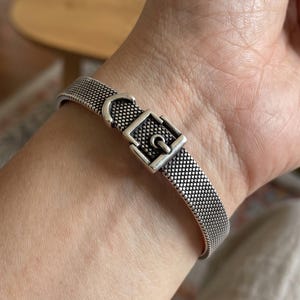 May include: A silver-toned metal bracelet featuring a textured, dotted surface pattern that mimics the appearance of a belt strap. The bracelet includes a central buckle mechanism with a prong and a rectangular frame, finished with an oxidized effect.