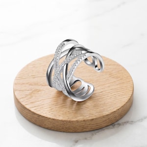 Könnte beinhalten: Ein silberfarbener Ring mit einem mehrreihigen Crossover-Design, bei dem zwei der Bänder mit kleinen klaren Steinen besetzt sind. Der Ring verfügt über eine offene Rückseite zur Größenanpassung und eine polierte metallische Oberfläche.