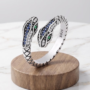 May include: A silver bypass ring shaped like a double-headed snake with a textured scale pattern. The snake heads are inlaid with rows of small blue and clear stones, accented by single green stones. The inner band is clearly stamped with S925.