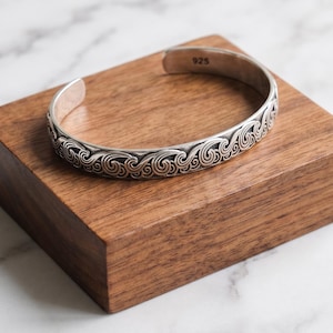 Pode incluir: Uma pulseira de prata tipo bracelete com um padrão de ondas em relevo que se repete ao longo da banda exterior. O interior da pulseira tem gravado o número 925, indicando o teor de prata. O metal tem um acabamento polido.