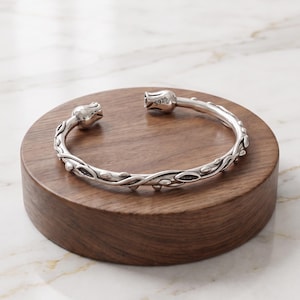 Può includere: Un bracciale rigido in argento con un motivo a vite intrecciata, piccoli dettagli a forma di foglia e due estremità a forma di tulipano. L'interno di una delle estremità a tulipano reca l'incisione S925, che indica la composizione in argento.
