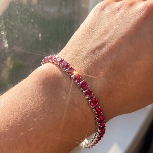 Könnte beinhalten: Ein Tennisarmband mit einer durchgehenden Reihe runder, leuchtend roter Steine, die in einen polierten, silberfarbenen Metallrahmen gefasst sind. Die Steine sind einheitlich in Größe und Form und reflektieren das Licht mit hellem Glanz.