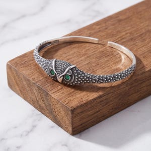 May include: A silver cuff bracelet featuring a detailed owl head design at the center. The owl has two small, vibrant green stones set as eyes. The band of the bracelet is textured with intricate feather-like engravings along the entire length.