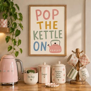 以下が含まれることがあります： ピンク、黄色、緑、青の文字で「POP THE KETTLE ON」と書かれ、ピンクのケトルのイラストが描かれた額入りアート。その下には、ピンクの電気ケトル、ティーバッグ用ボウル、ストライプ柄の砂糖・茶葉用キャニスター、4つのマグカップが掛かったマグツリーが並んでいる。
