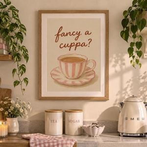 Könnte beinhalten: Ein gerahmter Kunstdruck mit einer rosa-weiß gestreiften Teetasse und Untertasse auf beigem Hintergrund. Der Text "fancy a cuppa?" steht in brauner Schreibschrift über der Illustration der Tasse. Der Rahmen ist aus hellem Holz.