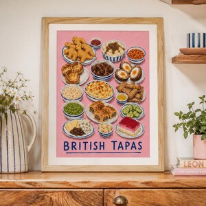 May include: A framed art print on a pink background featuring an illustration of various British food items in small bowls and plates, including nuggets, scotch eggs, and sausage rolls, with the text BRITISH TAPAS printed in blue at the bottom.