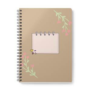 May include: A spiral-bound notebook with a tan cover featuring a central rectangular notepad graphic with a pink border and five silver rings. The cover is decorated with two light green leafy branches with small pink heart-shaped flowers.