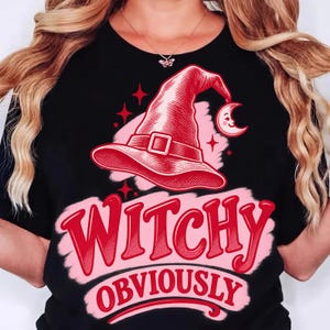 May include: A black t-shirt featuring a graphic of a red witch hat, a crescent moon with a face, and four-pointed stars. The text "WITCHY" and "OBVIOUSLY" is printed in red letters over a light pink brushstroke background on the front.