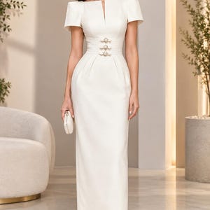 Peut inclure: Une robe longue couleur crème à manches courtes, dotée d'une encolure carrée avec une fente centrale et d'une taille ajustée ornée de trois boutons décoratifs. La jupe est droite et la tenue est associée à des mules blanches à bout pointu.