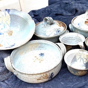 May include: A collection of handmade ceramic stoneware featuring a light blue and cream speckled glaze with brown leaf-like patterns. The set includes a large bowl, a lidded casserole dish, a smaller lidded pot, a handled bowl, and a small jar.