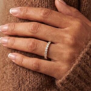 May include: A gold band ring featuring a continuous row of small, round, clear, faceted stones set along the entire circumference of the metal band. The ring is worn on the ring finger, showcasing a polished metallic finish and sparkling stones.
