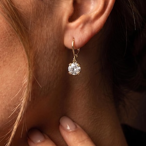 May include: A gold leverback earring featuring a single round, clear, faceted gemstone suspended from a delicate gold hook. The earring is worn in the earlobe, showcasing the metallic gold setting and the reflective quality of the stone.