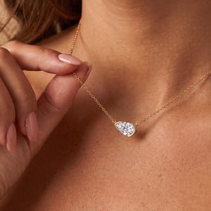 May include: A gold chain necklace featuring a single pear-shaped clear gemstone pendant in a bezel setting. The delicate metal chain connects to the sides of the teardrop-shaped stone, which rests against the collarbone area of the neck.
