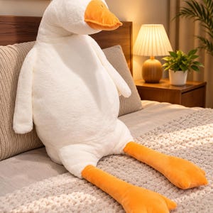 Pode incluir: Um grande peluche de ganso com pelo branco macio, um bico laranja brilhante e longas patas palmadas cor de laranja. O animal de peluche tem um corpo arredondado, olhos pretos pequenos e duas asas posicionadas nos lados.