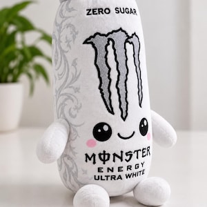 May include: A plush toy shaped like a white energy drink can with a grey top. It features the black Monster Energy logo, the text ZERO SUGAR, MONSTER ENERGY, and ULTRA WHITE. The can has a cute face with black eyes, a smile, and pink cheeks.