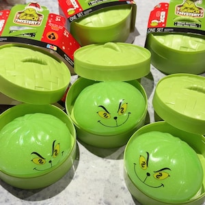 May include: A collection of Grinch Mystery Bun toy sets, each featuring a light green, round, lidded container resembling a bamboo steamer. Inside each container is a smooth, light green, spherical toy with a printed Grinch face and yellow eyes.