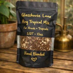 May include: A black stand-up pouch containing potting mix with yellow text that reads Glasshouse Lane Airy Tropical Mix For Aroids + Tropicals 1QT 10oz Hand Blended with a heart symbol. The pouch has a clear window showing the brown and white soil.