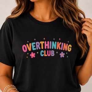 May include: A black short-sleeved t-shirt featuring the text OVERTHINKING CLUB in a multi-coloured, rounded font. The design includes small four-pointed star icons and two simple flower illustrations in pink and purple, arranged around the text.
