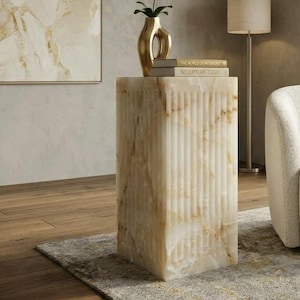May include: A rectangular fluted pedestal made of cream and beige veined stone. On top sits a gold-toned abstract vase containing a white orchid, and two books titled The World of Interiors and Sculpture Today.