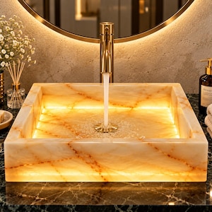 Könnte beinhalten: Ein quadratisches Waschbecken aus durchscheinendem Onyx-Stein mit warmen orangefarbenen und weißen Adern, das von innen beleuchtet wird. Ein goldfarbener Wasserhahn ist über dem Becken zentriert und lässt Wasser in den Abfluss fließen.