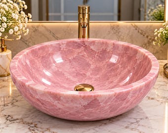 Puede incluir: Un lavabo de sobreponer redondo hecho de piedra rosa pulida con una textura veteada natural. El cuenco presenta un acabado liso y brillante con un desagüe dorado centrado, situado bajo un grifo a juego de color dorado.