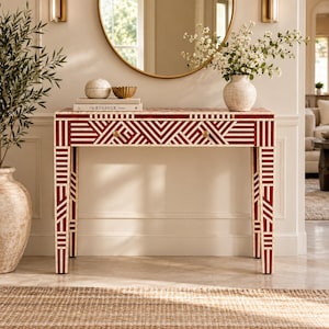 May include: A rectangular console table featuring a bold red and white geometric inlay pattern. The table has four straight legs and a single drawer with a small round knob. The surface holds a stack of books, a round decorative object, and a bowl.