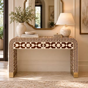 Puede incluir: Una mesa consola rectangular con marco curvo que presenta un patrón geométrico incrustado en marrón y crema. La mesa tiene dos cajones con pomos pequeños, patas de metal dorado y una superficie con un jarrón, libros, un cuenco y una lámpara.
