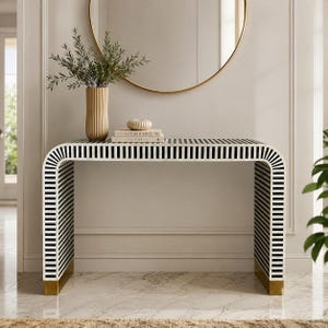 May include: A console table with a waterfall edge design featuring a black and white striped pattern. The table has a rectangular top and two side panels that curve downward to the floor, finished with gold-toned metal feet at the base.