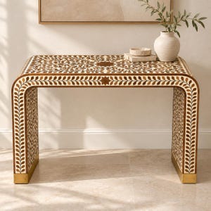 May include: A waterfall console table featuring an intricate brown and cream floral inlay pattern across the top and sides. The table has a rectangular silhouette with rounded edges and is finished with metallic gold-toned base accents.