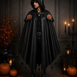 May include: A black hooded cape featuring orange jack-o'-lantern patches on the shoulders and dangling black charms along the hem. The cape is secured at the neck with a buckle strap and is worn over a black shirt and black trousers.